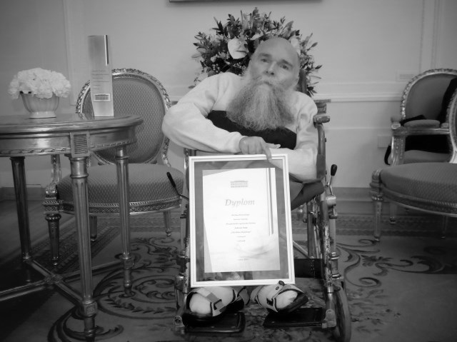 Czarno-białe zdjęcie. Centralnie na wózku Jan Arczewski w białej koszuli z długą siwą brodą, prezentuje na kolanach oprawiony dyplom.