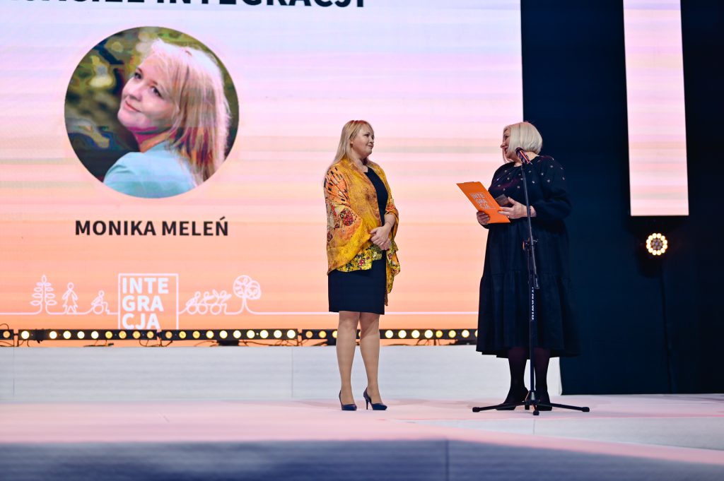 Scena 28.Wielkiej Gali Integracji. Na ogromnym ekranie zdjęcie laureatki Moniki Meleń. Na scenie, przed ekranem dwie osoby – laureatka i Ewa Pawłowska, moment wręczenia medalu.