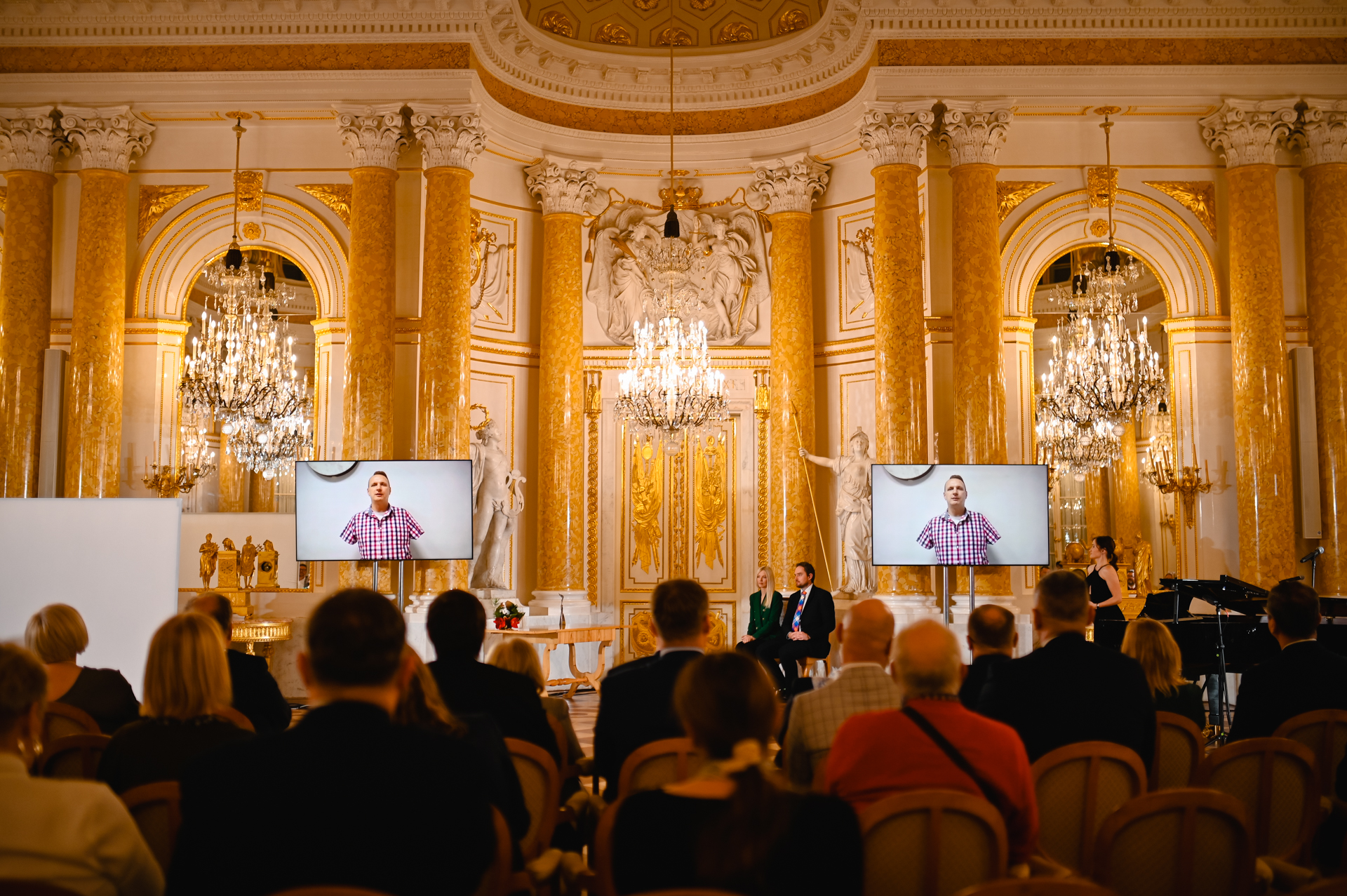 Reprezntacyjna sala Zamku Królewskiego ze złoconymi sztukateriami, na scenie, po o bu stronach 2 bilbordy z wizerunkiem laureata Przemysława Wieczorka.