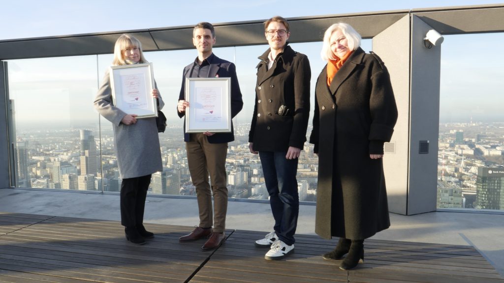 cztery osoby stoją na dachu wieżowca, dwie z nich prezentują oprawione w ramki certyfikaty