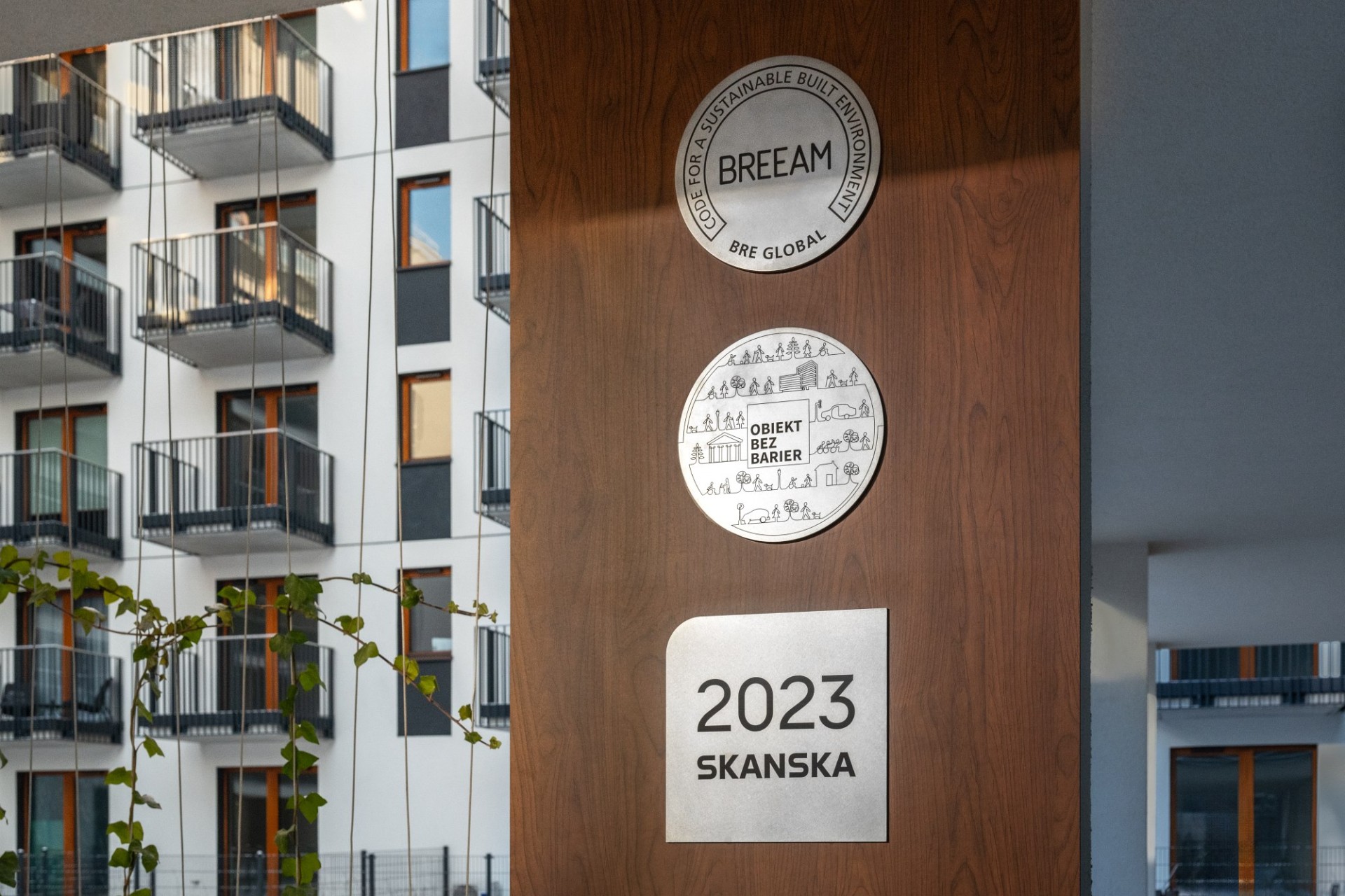 Drewniany fragment elewacji ze srebrnymi tablicami BREEAM, Obiekt bez barier, 2023 SKANSKA, w głębi elewacja apartamentowca z balkonami.
